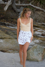 Woman in a striped swimwear one piece and white towel skort with brown shell design standing against a rocky background